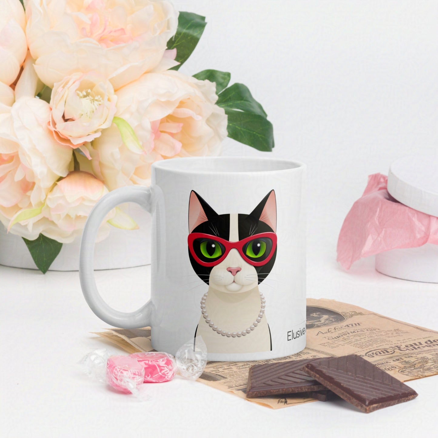 Mug with a cat design wearing sunglasses, placed on a surface with flowers and chocolate.